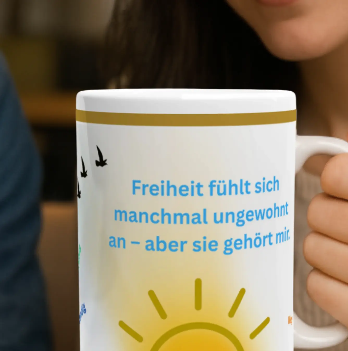 Eine Person hält eine Tasse mit dem Text 'Freiheit fühlt sich manchmal ungewohnt an - aber sie gehört mir.' Die Tasse zeigt eine Sonne und Vögel.